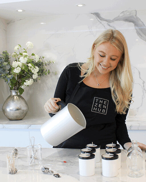 blonde haired women with THE ZEN HUB apron on pouring candles in kitchen with white marble backboard