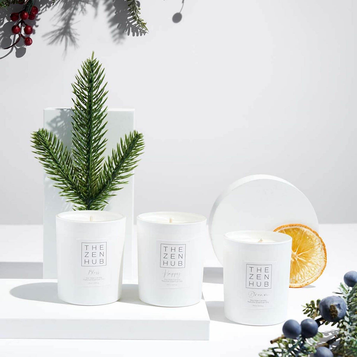 3 travel candles on white surface with white background. There are berries, Christmas tree sprigs and dried orange segments as Christmas decorations