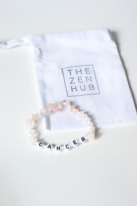 rose quartz crystal healing bracelet with lettered beads spelling cancer. Crystal bracelet is on white cotton branded bag and surface. 